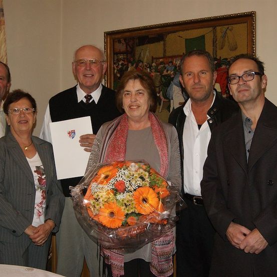 Eine Gruppe von Menschen posiert für ein Foto, eine Frau hält einen Blumenstrauß, eine andere ein Zertifikat. Sie stehen vor einem Gemälde an der Wand.