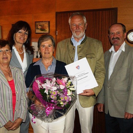Eine Gruppe von fünf Personen steht in einem Raum. Eine Frau hält einen Blumenstrauß und ein Zertifikat. Der Mann auf der rechten Seite hält ein Zertifikat. Der Mann in der Mitte trägt eine grüne Jacke. Eine Uhr ist an der Wand.