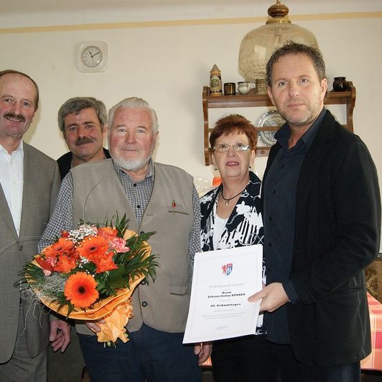 Eine Gruppe von Menschen steht zusammen, ein Mann hält einen Blumenstrauß und ein Zertifikat, während andere zusehen.