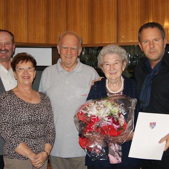 Fünf Personen stehen in einem Raum zusammen, wobei eine ältere Frau einen Blumenstrauß und ein Zertifikat hält. Hinter ihr hält ein Mann ein Zertifikat. Die anderen drei Personen lächeln und schauen in die Kamera.