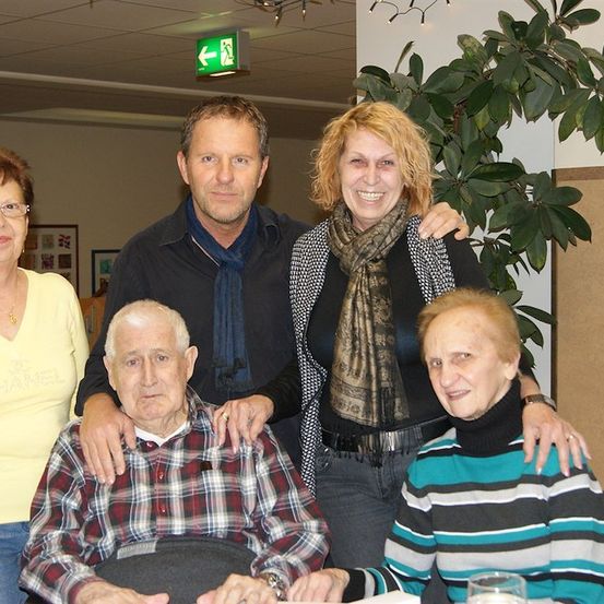 Eine Gruppe von Menschen, die für ein Foto zusammenstehen, darunter ein älterer Mann, der auf einem Stuhl sitzt, eine Frau mit Brille und ein Mann mit einem Schal.