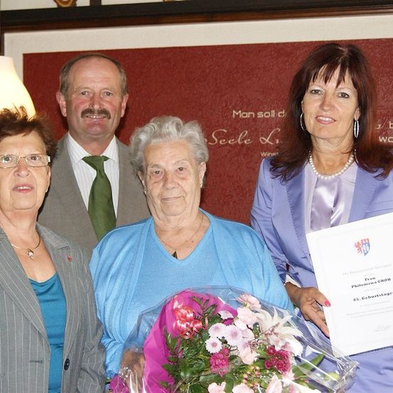 Vier Menschen stehen zusammen und lächeln. Die Frau in der Mitte hält einen Blumenstrauß und ein Zertifikat. Hinter ihr hält ein Mann ein Zertifikat. Die Frau auf der linken Seite trägt eine Brille und eine Halskette. Die Frau auf der rechten Seite trägt eine Halskette und Ohrringe. Eine rote Wand mit Text steht hinter ihnen.