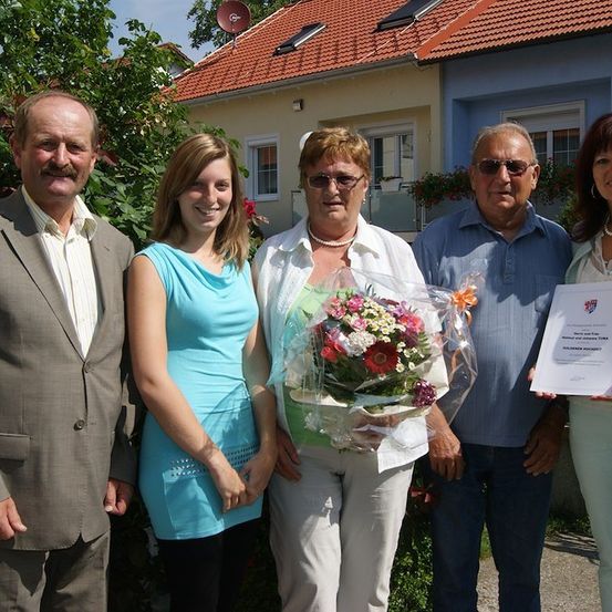 Fünf Personen stehen vor einem Haus, eine Frau hält einen Blumenstrauß und eine andere ein Zertifikat. Die Frau in der Mitte trägt eine Brille und eine Halskette. Dahinter stehen Pflanzen und ein Haus mit rotem Dach und Solarpanels.