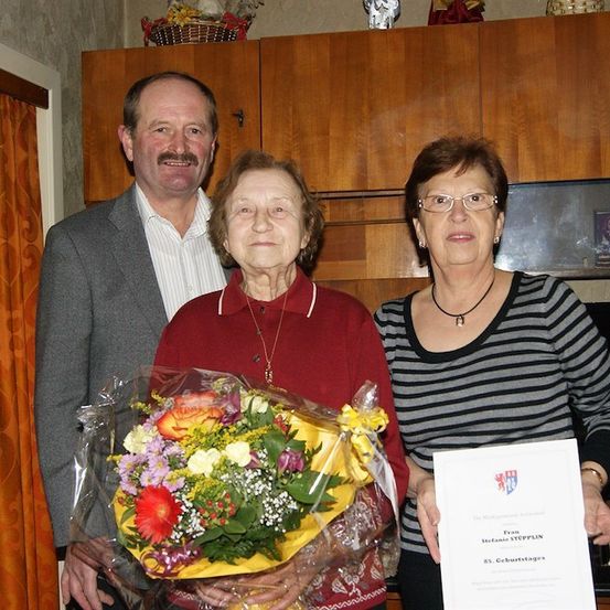 Drei Personen stehen in einem Raum, eine hält einen Blumenstrauß und eine andere ein Zertifikat. Die Frau in der Mitte trägt eine Halskette und der Mann links hat einen Schnurrbart. Hinter ihnen steht ein Holzschrank mit einem Korb obenauf.