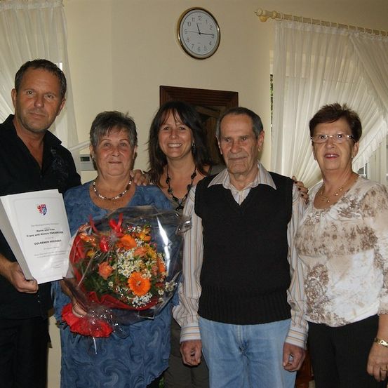 Eine Gruppe von Menschen steht zusammen und lächelt für ein Foto. Der Mann auf der linken Seite hält ein Zertifikat und einen Blumenstrauß. Die Frau in der Mitte hält einen Blumenstrauß. Der Mann auf der rechten Seite trägt eine Brille. Hinter ihnen ist eine Wand mit einer Uhr und einem Fenster mit weißen Vorhängen.