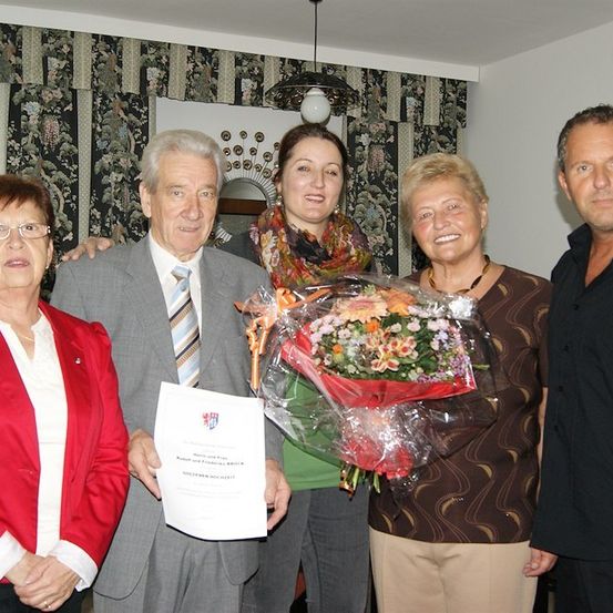 Fünf Menschen stehen lächelnd in einem Raum. Der Mann in der Mitte hält ein Zertifikat und einen Blumenstrauß. Die Frau rechts hält den Strauß. Die Frau links trägt eine Brille und einen roten Blazer. Die Frau in der Mitte trägt einen Schal. Der Mann rechts trägt ein schwarzes Hemd.