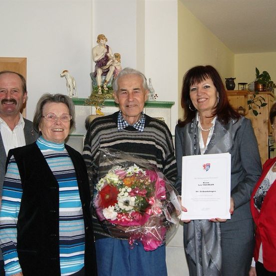 Eine Gruppe von Menschen steht in einem Raum zusammen, ein Mann hält einen Blumenstrauß und ein Zertifikat, während andere lächeln und in die Kamera schauen.