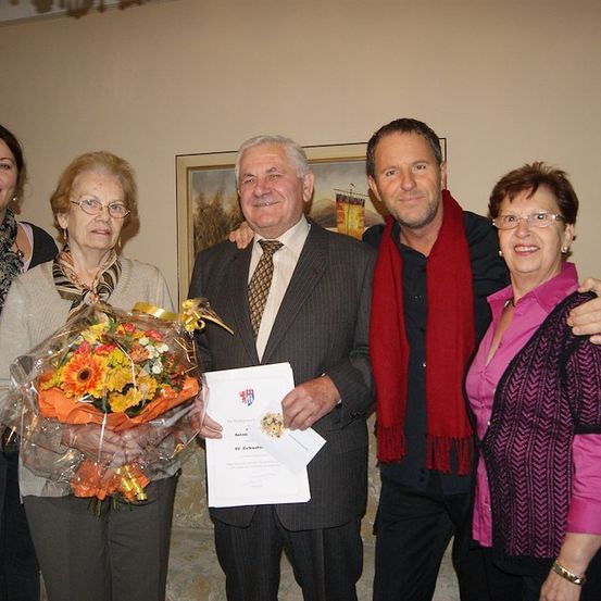 Eine Gruppe von Menschen steht zusammen und lächelt für ein Foto. Ein Mann in Anzug hält ein Zertifikat und einen Blumenstrauß. Eine Frau mit Brille hält den Strauß.