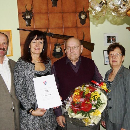 Eine Gruppe von Menschen steht lächelnd für ein Foto zusammen. Der Mann in der Mitte hält ein Zertifikat und einen Blumenstrauß. Hinter ihnen ist eine Wand mit aufgehängten Hirschköpfen und einer Kronleuchter zu sehen.