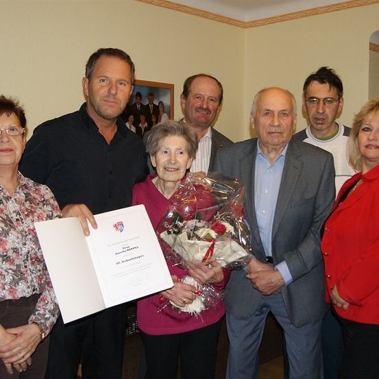 Eine Gruppe von Menschen steht zusammen und lächelt, wobei eine Person einen Blumenstrauß und ein Zertifikat hält. Die Frau in der Mitte hält den Strauß mit beiden Händen.