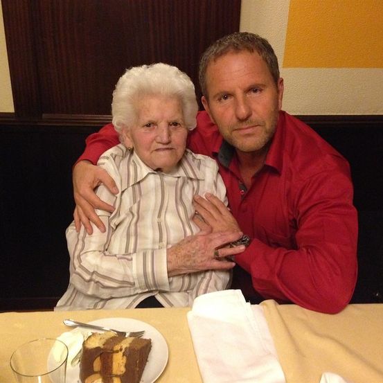 Eine ältere Frau und ein Mann sitzen zusammen an einem Tisch, wobei der Mann seinen Arm um ihre Schultern gelegt hat. Sie lächeln beide und schauen in die Kamera. Vor ihnen steht ein Stück Kuchen auf einem Teller.