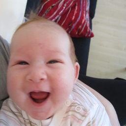 Ein Baby mit glücklichem Gesichtsausdruck wird von jemandem gehalten, der ein rot-blau gemustertes Shirt trägt.
