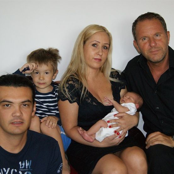 Ein Familienfoto mit einem Mann, einer Frau und zwei Kindern. Die Frau hält ein Baby, während der Mann neben ihr sitzt. Die Kinder sitzen auf dem Schoß der Frau und auf dem des Mannes.