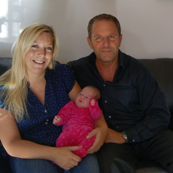 Eine lächelnde Frau hält ein Baby in rosa Schlafanzug, während sie auf einer Couch mit einem Mann sitzt. Sie alle schauen in die Kamera.