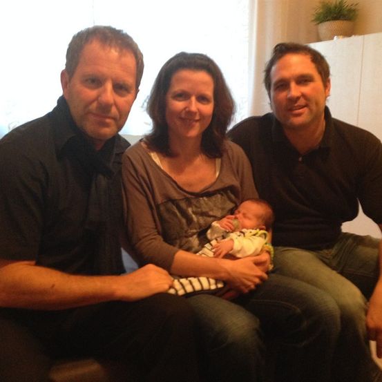 Drei Erwachsene und ein Baby sitzen auf einer Couch und lächeln für ein Foto. Das Baby wird von einer Frau gehalten.