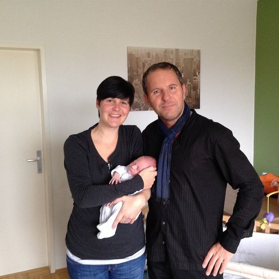 Ein Mann und eine Frau stehen in einem Zimmer und halten ein Neugeborenes, während sie lächeln. Der Raum hat weiße Wände und eine grüne Akzentwand.