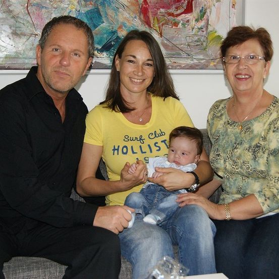 Eine Familie mit drei Personen, bestehend aus einem lächelnden Mann, einer Frau und einem Baby, sitzt auf einer Couch mit einem farbenfrohen Gemälde im Hintergrund.