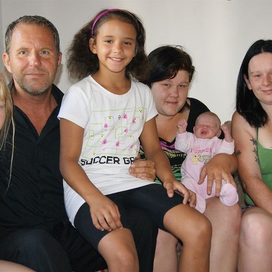 Eine Familie mit fünf Personen posiert für ein Foto. Der Mann trägt ein schwarzes Hemd, und das Mädchen in der Mitte trägt ein weißes T-Shirt mit einem Fußball-Grafik. Die Frau auf der rechten Seite hält ein Baby.