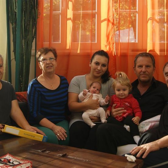 Eine Familie mit fünf Personen, darunter zwei kleine Kinder, sitzt auf einem Sofa im Wohnzimmer mit orangefarbenen Vorhängen. Die Frau in der Mitte hält ein Baby, während die andere Frau ein Kleinkind hält.