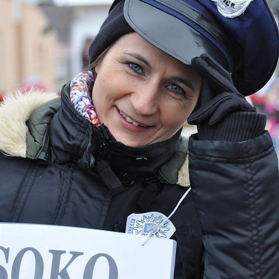 Eine Frau in einer Polizeiuniform, lächelnd und ein Schild mit der Aufschrift 'SOKO' haltend. Sie trägt einen Hut und Handschuhe.