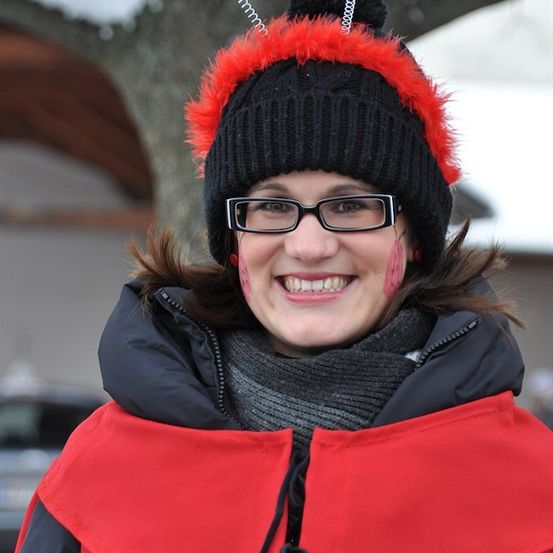Eine Frau trägt einen roten und schwarzen Winterhut mit rotem Pelzbesatz, eine Brille und eine rote und schwarze Jacke mit Kapuze. Sie hat rotes Gesichtsmal.