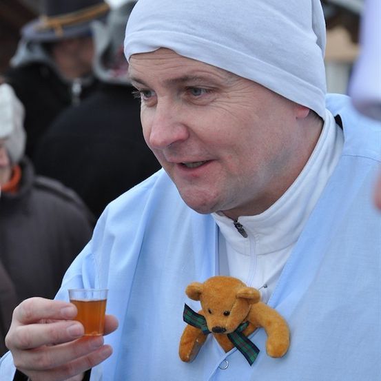 Ein Mann in einem blauen Gewand und einer weißen Kopfbedeckung hält ein Glas Bier in der Hand, mit einem kleinen Teddybär an seiner Brust befestigt.