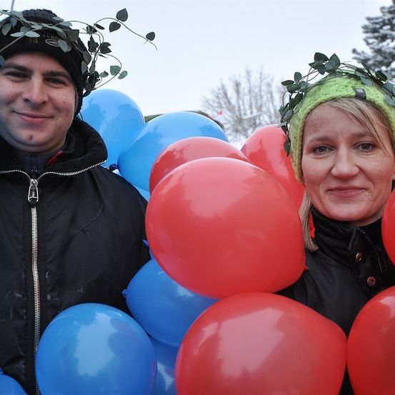 Zwei Personen stehen umgeben von Luftballons und tragen Kränze auf dem Kopf. Der Mann trägt eine schwarze Jacke und die Frau eine grüne Mütze.