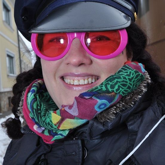 Eine Frau mit schwarzem Hut, rosa Sonnenbrille und buntem Schal lächelt vor einem Gebäude.