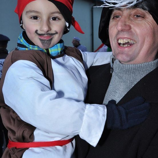 Ein junger Junge, der einen Hut mit einer roten Bandana und einem falschen Schnurrbart trägt, umarmt einen älteren Mann in einem schwarzen Mantel und Hut mit Federn.