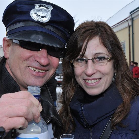 Ein Mann mit Polizeimütze und Sonnenbrille hält eine Flasche und lächelt. Daneben lächelt eine Frau mit Brille und blauer Jacke in die Kamera.