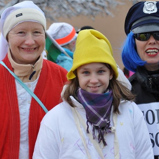 Drei Frauen in Kostümen: eine Nonne, ein Kind mit gelber Mütze und eine Polizistin mit blauen Haaren. Sie lächeln und stehen zusammen.