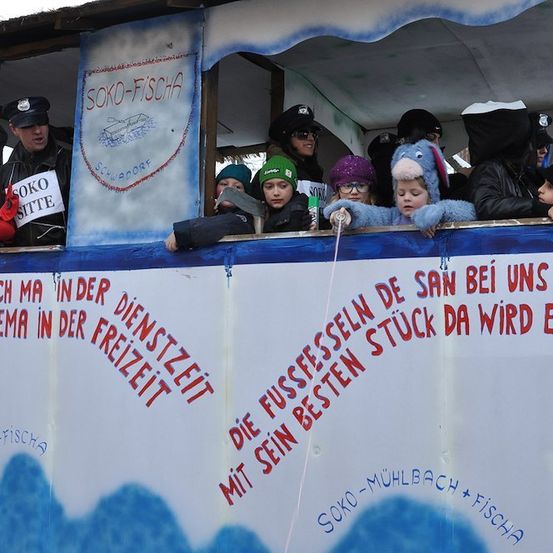 Eine Gruppe von Menschen in Winterkleidung schaut aus einem dekorierten Wagen heraus, mit einem Banner, das 'SOKO-FISCHA' und einen Slogan auf Deutsch liest. Ein Mann hält ein Schild mit der Aufschrift 'SOKO SITTE'.