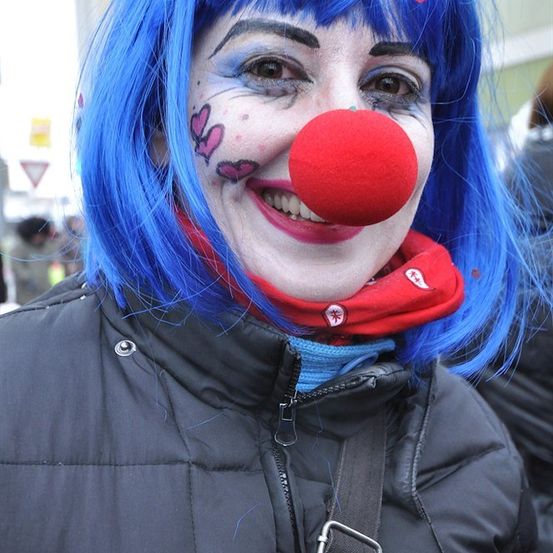 Eine Frau mit blauen Haaren, rotem Nasenbalg und Gesichtsbemalung, lächelt in einer schwarzen Jacke.