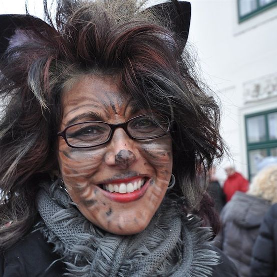 Eine Frau, die als Katze verkleidet ist, mit Brille und Schal, mit Make-up im Gesicht, lächelt vor einem Gebäude.