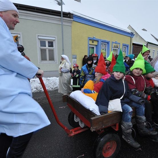 Ein Mann in einem weißen Gewand zieht einen Holzwagen mit Kindern in farbenfrohen Elfenhüten. Sie sind auf einer verschneiten Straße mit Gebäuden im Hintergrund.