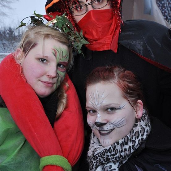Drei junge Frauen in Kostümen, eine mit grüner Gesichtsbemalung, eine mit Katzengesicht und eine mit roter Maske und Hut.