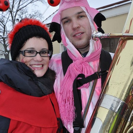 Ein Mann in einem rosa Schweinekostüm und eine Frau in einem roten Mantel lächeln und posieren für ein Foto mit einem Tuba im Vordergrund.