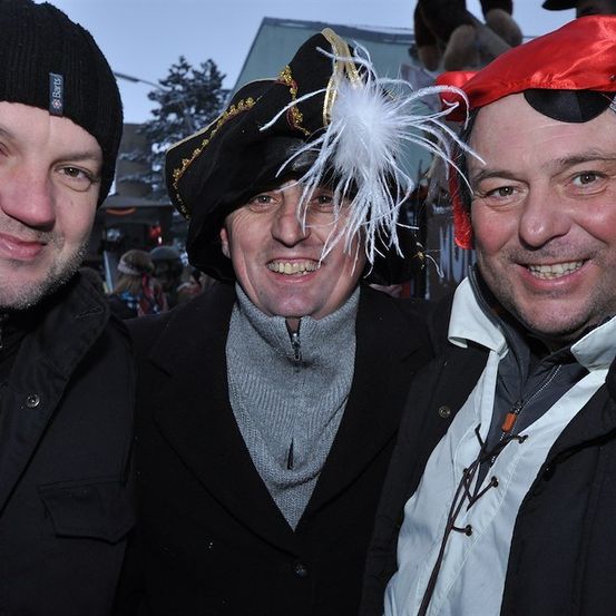 Drei Männer in Winterkleidung und festlichen Hüten, lächeln für ein Foto. Der Mann in der Mitte trägt einen Piratenhut mit einer Feder, während die anderen Beanie-Hüte tragen.
