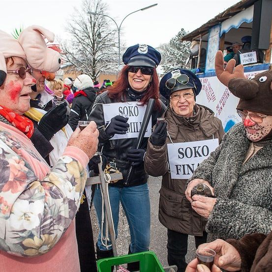 Eine Gruppe von Menschen in Kostümen, darunter ein Schwein, Polizisten und ein Rentier, stehen in einem Außenbereich, wahrscheinlich auf einem Festival. Sie halten Schilder und trinken aus Tassen. Hinter ihnen steht ein Gebäude mit einem Schild und einem Baum.