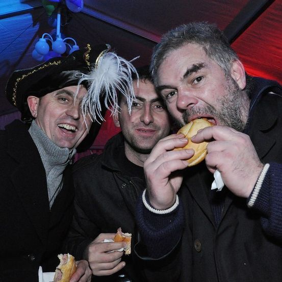 Drei Männer in Winterkleidung und Partyhüten essen Hot Dogs auf einer Party.