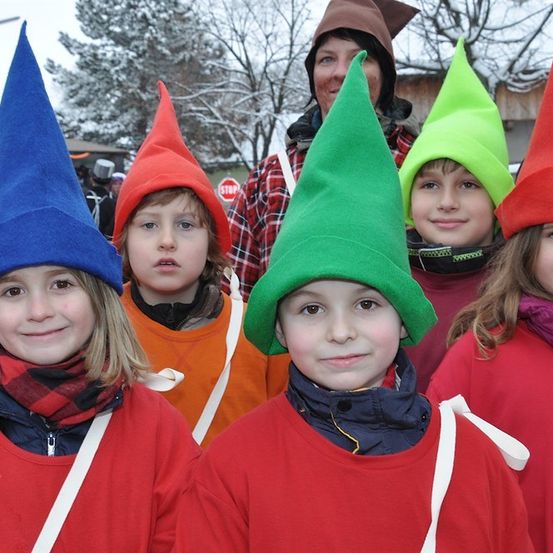 Eine Gruppe von Kindern in roten Kostümen mit Kegelhüten in verschiedenen Farben posiert für ein Foto in einer verschneiten Außenumgebung.