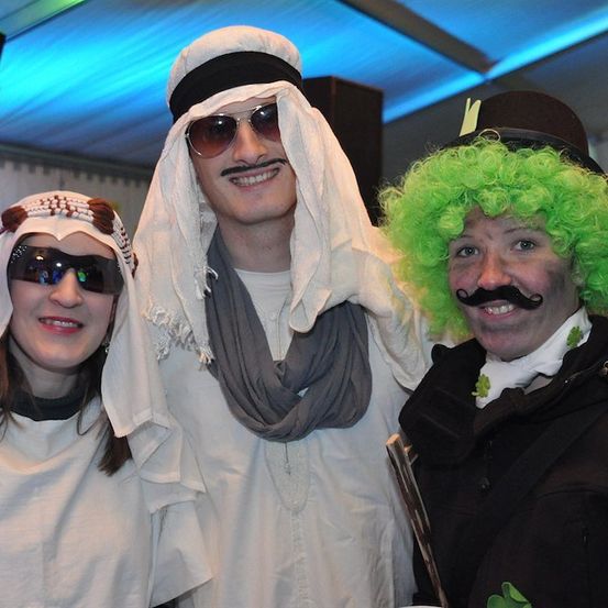Drei Menschen in Kostümen posieren für ein Foto. Eine Frau in einem weißen Gewand, ein Mann in einem weißen Gewand mit Schal und ein Mann mit grüner Perücke und Schnurrbart.