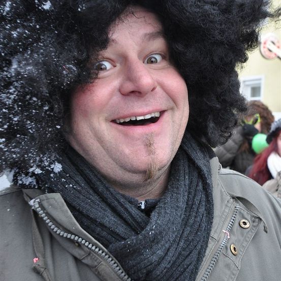 Ein Mann mit Afro-Mütze und Schal lächelt für die Kamera, Schneeflocken auf seinem Haar, mit einem verschwommenen Hintergrund von Menschen und Gebäuden.