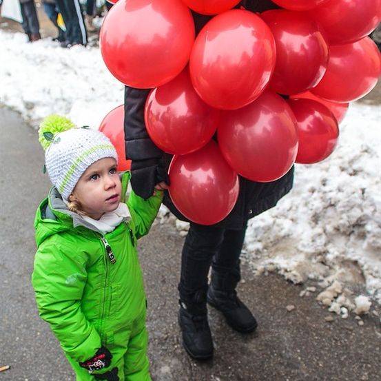 Ein kleines Kind in einem grünen Schneeanzug hält eine Menge roter Luftballons in der Hand, während es auf einer verschneiten Straße steht.