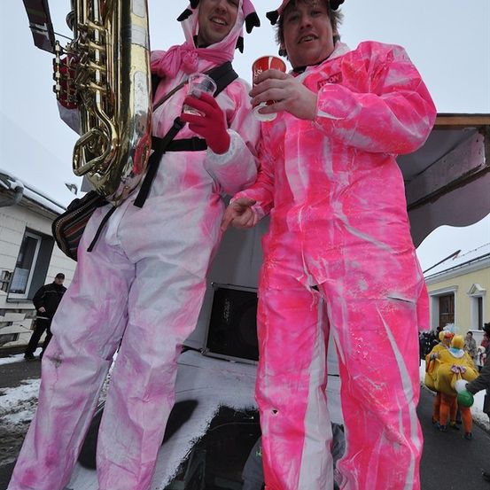 Zwei Männer in rosa Kostümen mit Schweinemasken, einer hält einen Becher und der andere eine Sousaphon, stehen vor einem Gebäude mit verschneiten Hintergrund.