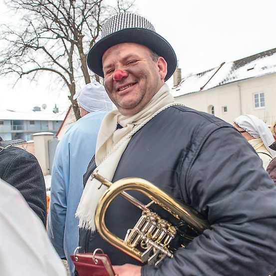 Ein Mann mit roter Nase und schwarzem Hut lächelt und hält eine Trompete in einer verschneiten Umgebung im Freien.