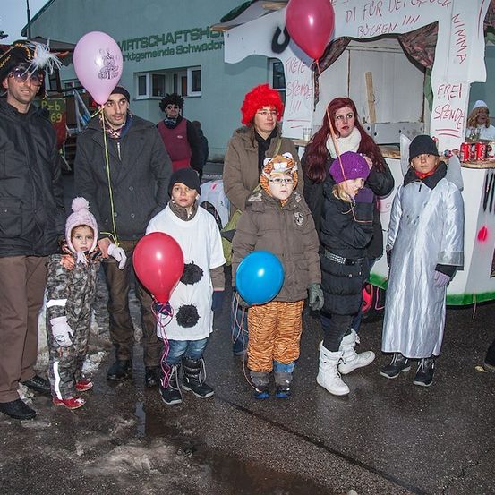 Eine Gruppe von Menschen in verschiedenen Kostümen, darunter ein Pirat, eine Kuh und ein Tiger, steht vor einem dekorierten Stand mit Luftballons und einem Schild, das 'Frei Spende' steht.