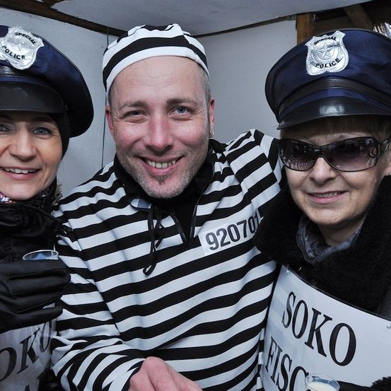 Drei Personen, die als Polizisten und ein Gefangener gekleidet sind, posieren für ein Foto. Der Mann in der Mitte trägt ein gestreiftes Häftlingsuniform mit der Nummer 920701. Die Frau links trägt einen Polizeihut und Handschuhe. Die Frau rechts hält ein Schild mit der Aufschrift 'SOKO'.