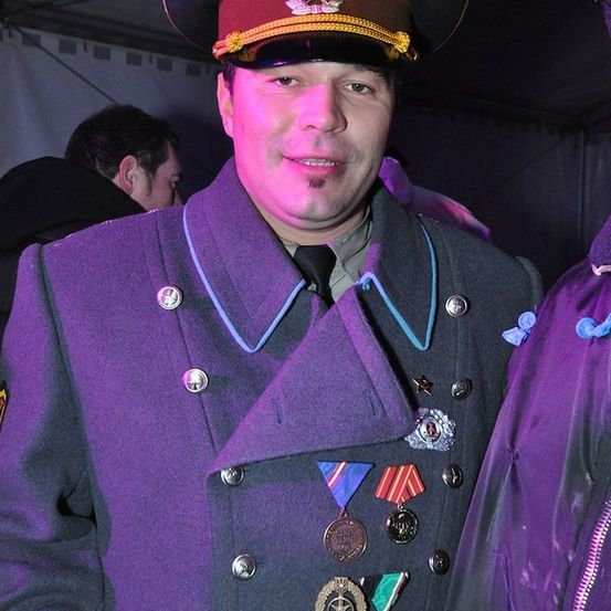Ein Mann in einem lila Militäruniform mit einem roten Stern auf seinem Hut und mehreren Medaillen auf seiner Brust. Er lächelt und schaut in die Kamera.