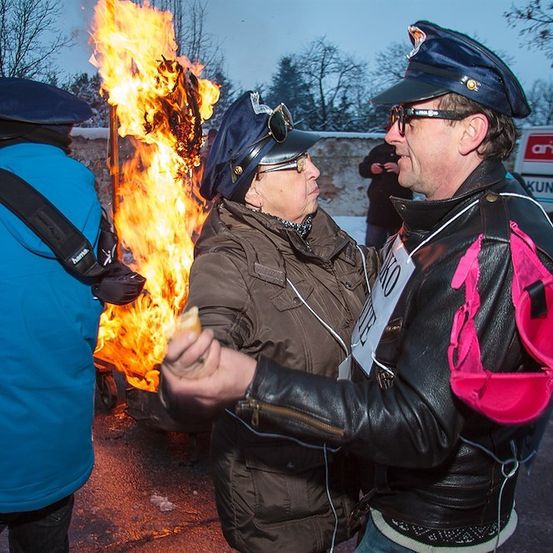 Ein Mann und eine Frau in Hüten und Lederjacken stehen vor einem Lagerfeuer, wobei ein Mann ein Sandwich hält und ein Mann mit einem Rucksack hinter ihnen steht.
